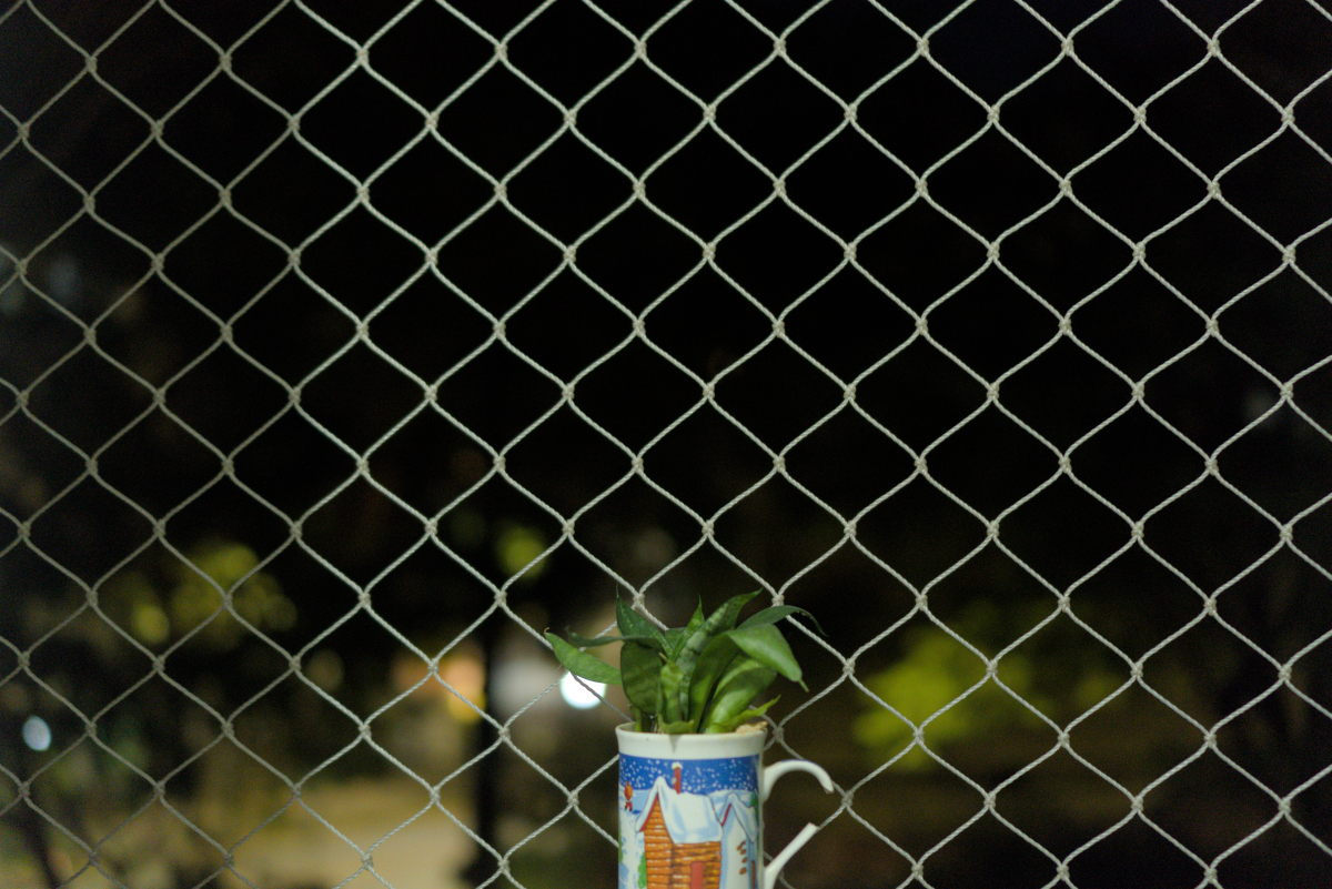 Ao centro, na parte de baixo, uma caneca com a asa quebrada e uma ilustração natalina. Atrás uma rede de proteção, possivelmente de uma janela. Ao fundo, borrado, as luzes noturnas da cidade.